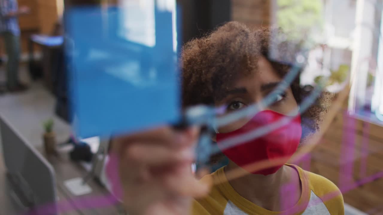 African american woman wearing face mask writing on glass board at modern office