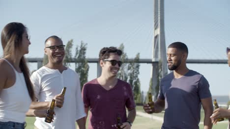 smiling young people clinking beer bottles in park.