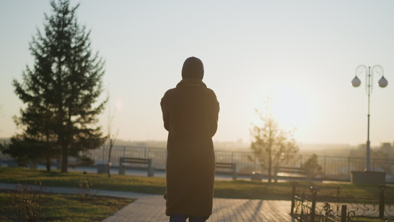 일몰에 공원을 가로질러 천천히 그리고 슬프게 걸어가는 여자의 뒷면 사진. 그녀의 실루은 황금 시간 빛에 의해 부드럽게 조명되어 평온하지만 우울한 분위기를 만니다.