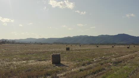 Fly-over-haystacks-in-the-countryside-field-with-mountain-ridge-in-the-background-on-a-bright,-clear,-sunny-day