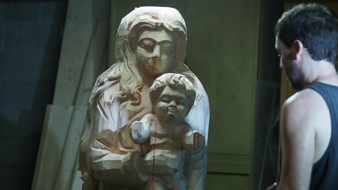 Sculptor Carefully Examines A Partially Finished Wooden Religious Statue Of Mary And Baby Jesus In His Workshop Setting