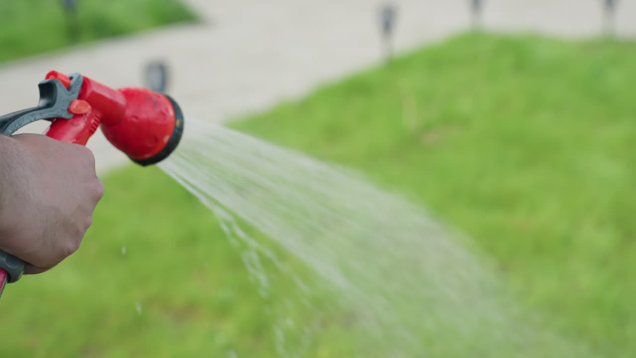 close up of hairy hand holding red hose nozzle gently watering lush grass in sunny backyard, water spray shimmering on vibrant green blades, smooth blur background
