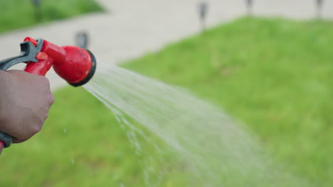 close up of hairy hand holding red hose nozzle gently watering lush grass in sunny backyard, water spray shimmering on vibrant green blades, smooth blur background