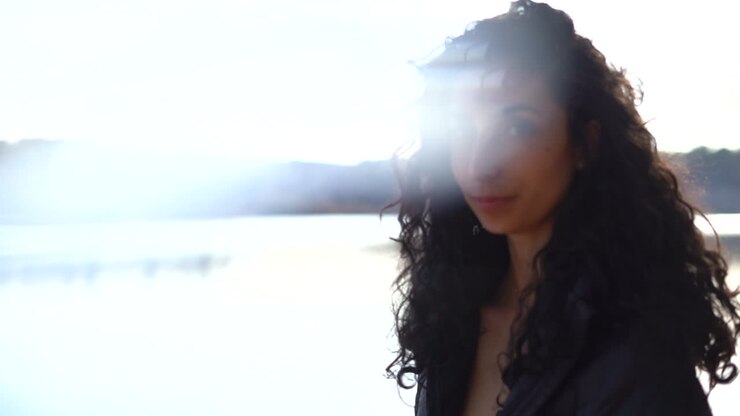 Pretty, young woman with thick, long, curly, brunette hair at a lake by the forest and the bright sunshine behind her - orbit shot with light leak, lens flare