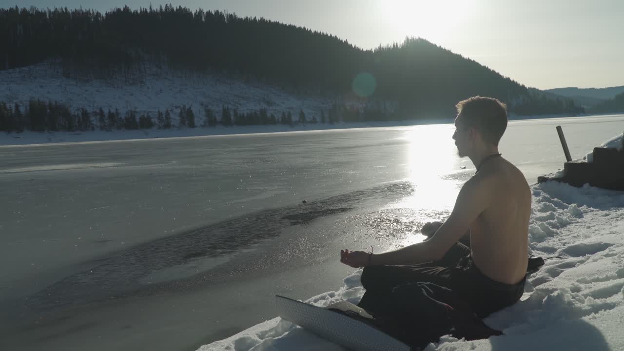 hombre meditando en una alfombra sentado en la nieve junto a un lago de montaña congelado con sol en el horizonte y luz del sol brillando en la superficie cubierta de hielo