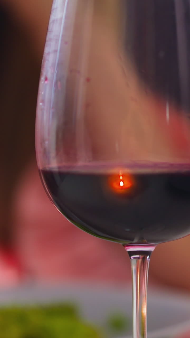 Wineglass of delicious collectible dark red wine on table against blurred view of woman eating salad in restaurant extreme closeup slow motion