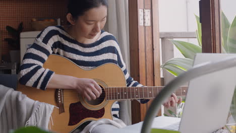 Junge-Asiatische-Frau,-Die-Zu-Hause-Gitarre-Spielt,-Angehende-Musikerin,-Die-Mit-Einem-Laptop-Das-Spielen-Eines-Musikinstruments-Lernt-Und-Dabei-Den-Kreativen-Ausdruck-Beim-Üben-Von-Musik-Genießt