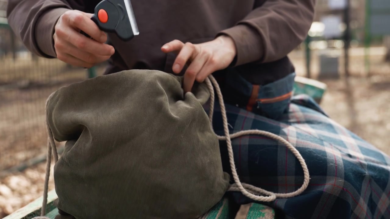 Una persona siede all'aperto su una panchina, maneggiando una borsa a sacchetto verde oliva con coulisse e una texture del cordino visibile. Un cane appare a intermittenza nell'inquadratura mentre le mani interagiscono con oggetti all'interno o vicino alla borsa.