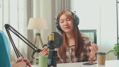 over shoulder view, young asian woman in headphones singing into microphone while recording podcast with male host in studio