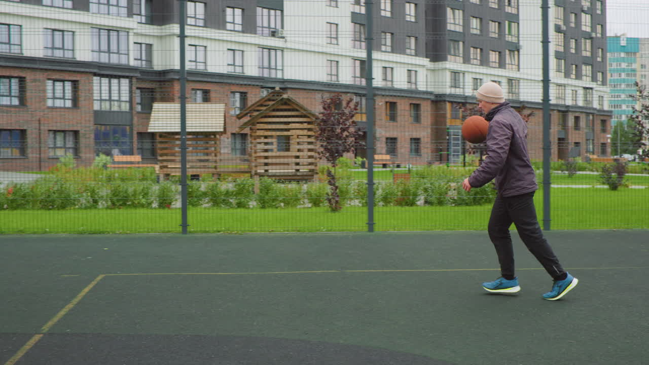 Un uomo anziano di etnia caucasica gioca a basket all'aperto in un campo urbano recintato, palleggia lungo il perimetro, avanza verso il canestro per un layup, indossa un piumino e scarpe da ginnastica, sullo sfondo un moderno appartamento, cielo nuvoloso