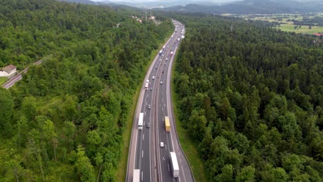 drone closing-in on traffic on the two-lane highway surrounded with green tall trees overpass and power lines beside a rail-system