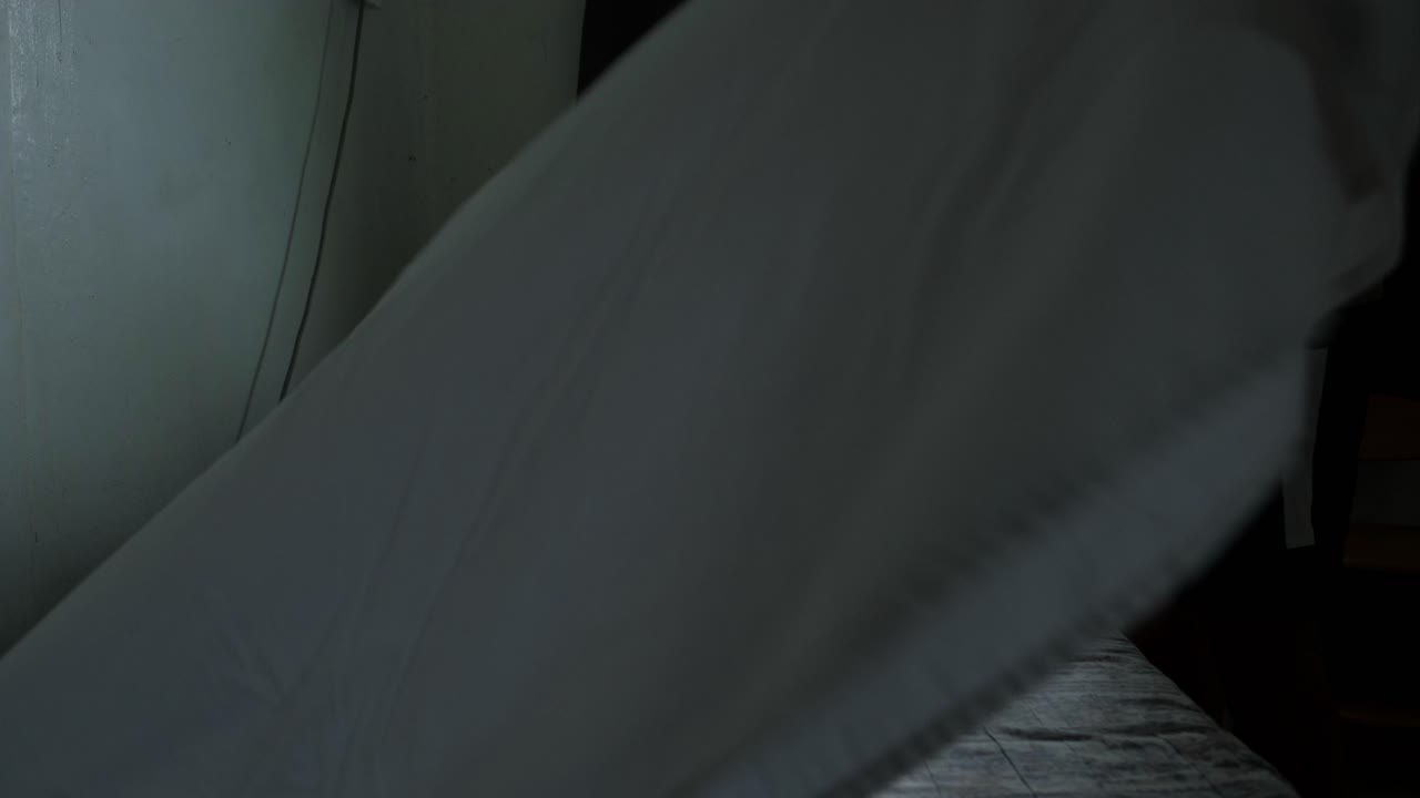 Static shot of young man arranging bedsheets and making bed in sunlit bedroom.