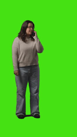 Vertical-Video-Full-Length-Shot-Of-Young-Woman-Talking-On-Mobile-Phone-Standing-Against-Green-Screen