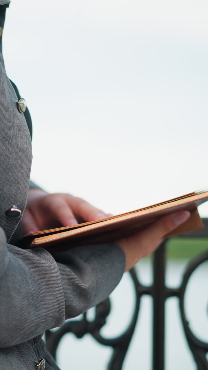 vista parcial de cerca de una mujer sosteniendo un libro abierto con su mano derecha descansando en las páginas y la mano izquierda apoyando debajo de ella, vestida de gris, de pie contra una valla de hierro, leyendo con atención