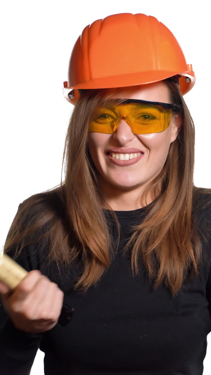 A beautiful woman in orange eyeglasses and helmet is holding a hammer in her hands and hitting herself by it in her hand on a white background in the studio. Engineer Vertical video