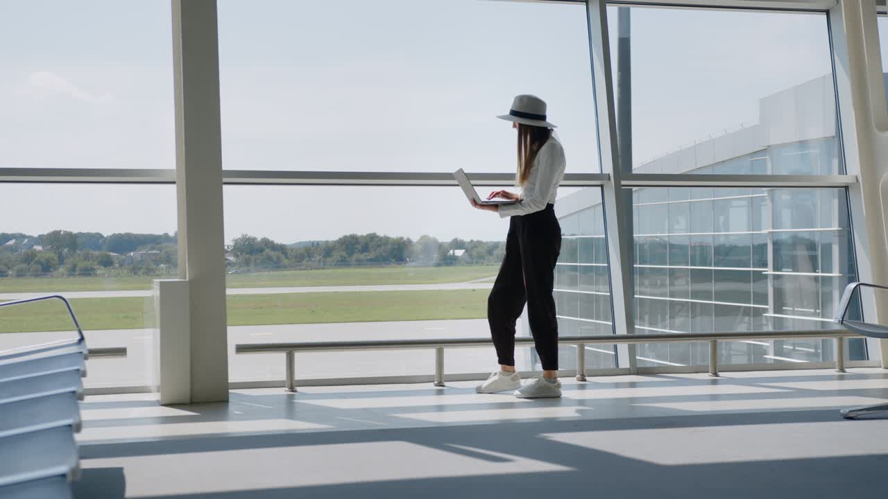 Young pretty woman printing on laptop at airport. Tourist freelancer works and waits for flight in waiting room. Concept remote work. Silhouette against the background of a large window. work remotely