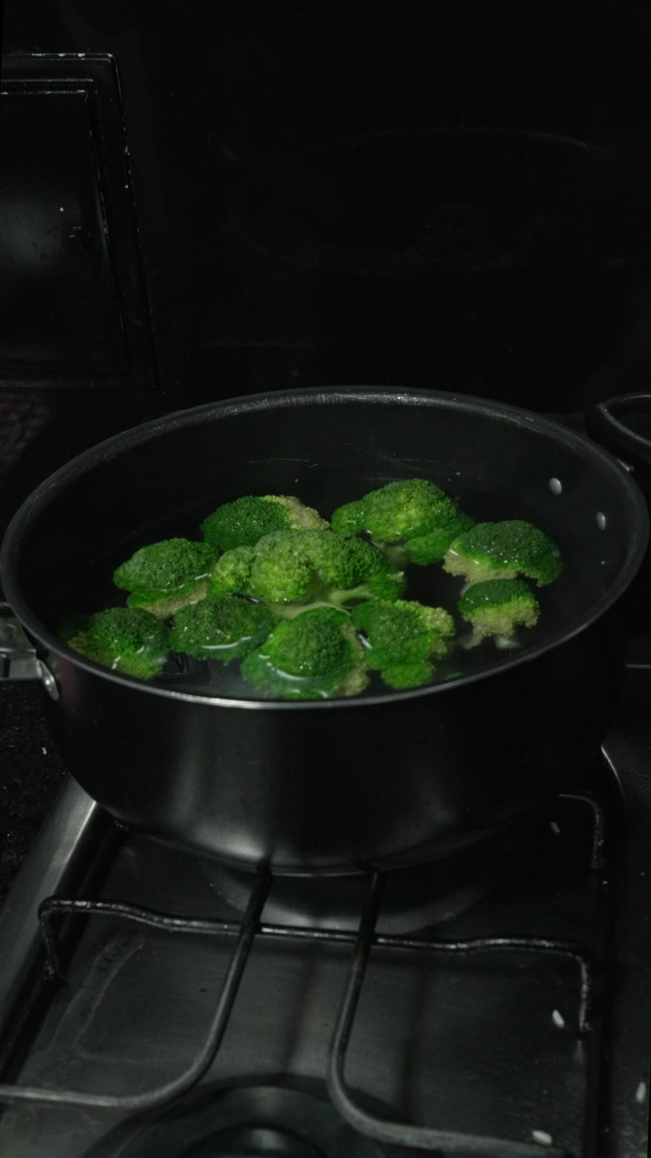 Pezzi di broccoli sobbollono in una pentola nera di acqua calda su un fornello a gas in acciaio inossidabile in una cucina domestica buia, utile per la cucina sana o per immagini di ricette