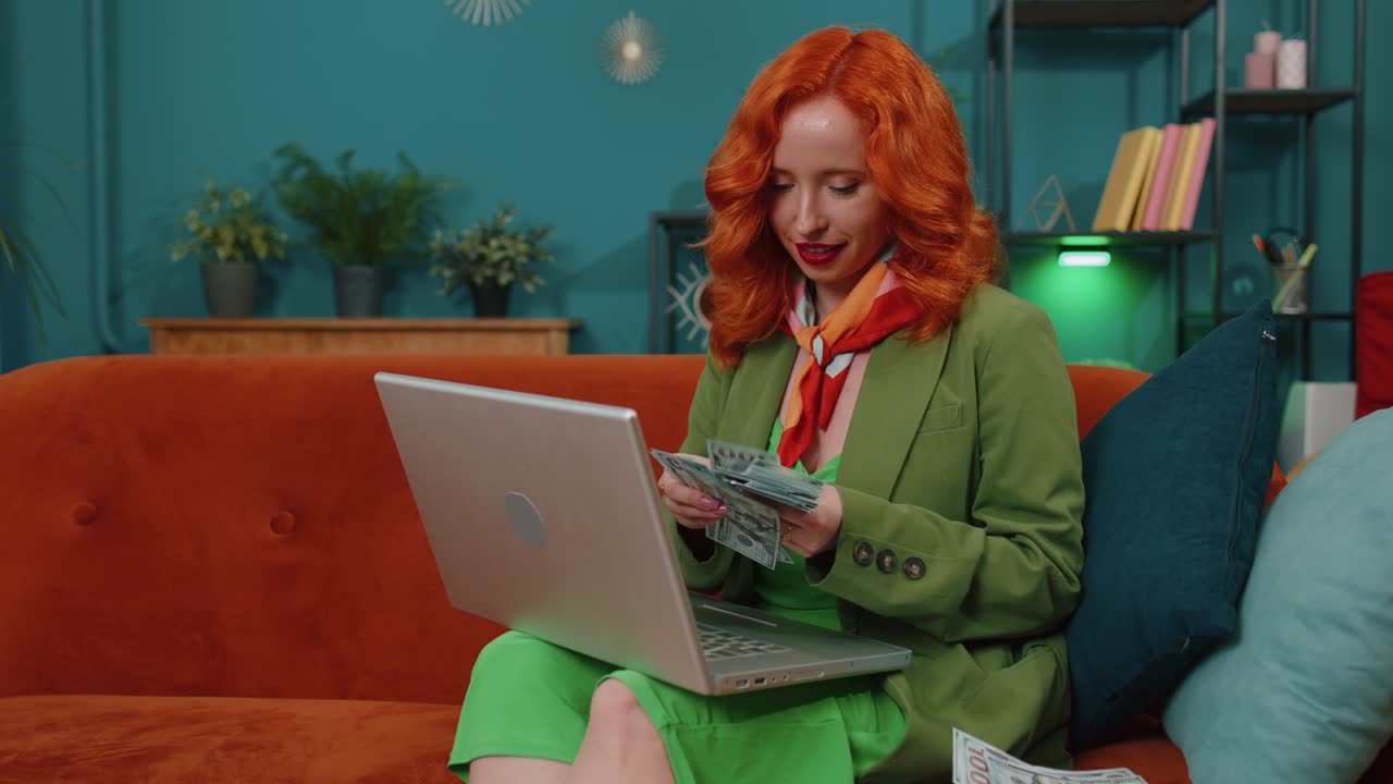 улыбающаяся счастливая женщина, считающая деньги, наличные и использующая ноутбук, вычисляет доходы дома.