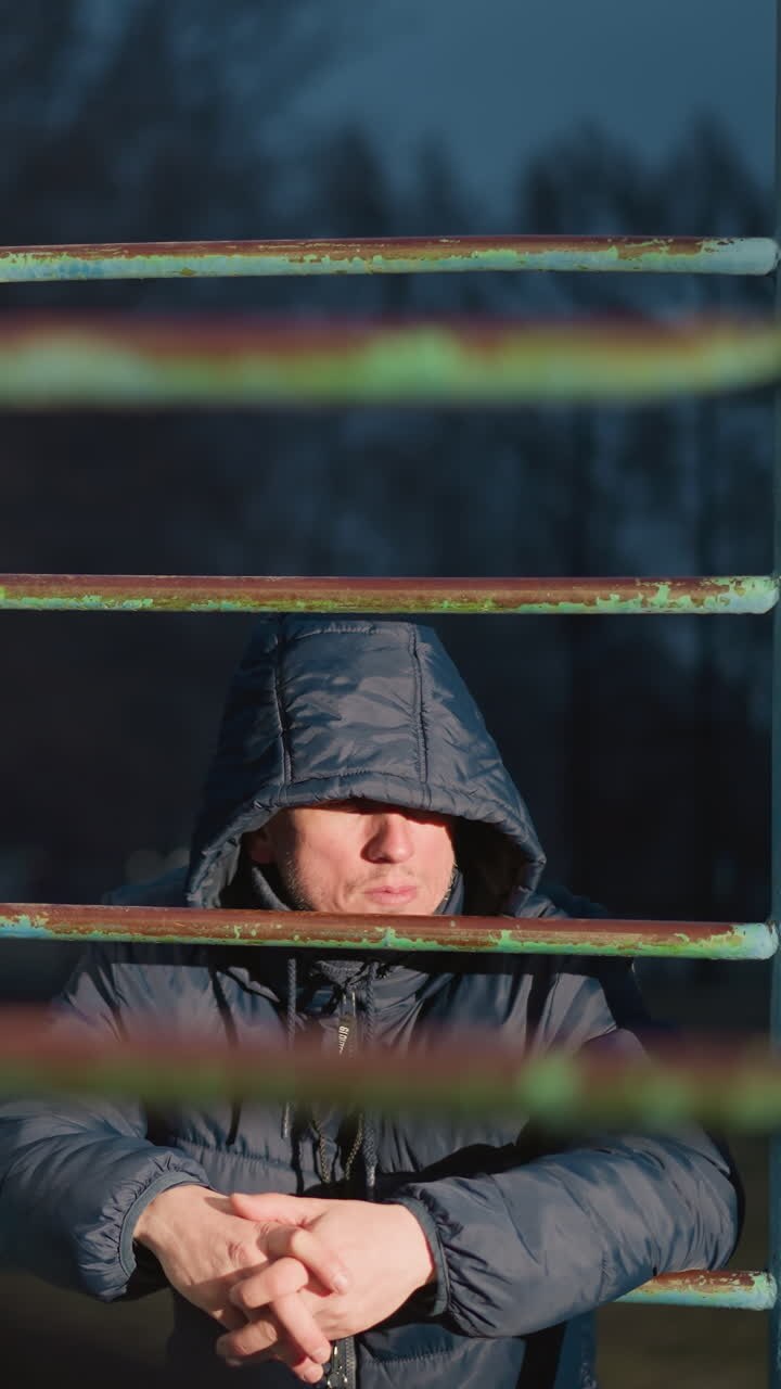 Close-up of two individuals resting on an iron bar, with one in a hooded jacket looking away as light reflects off him, the background features a blurred building and a moving car