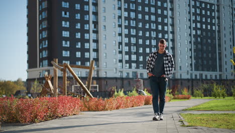 white man in checkered shirt walks along paved park path with hands in pockets during sunny day as distant pedestrian moves near modern residential buildings surrounded by greenery and red shrubs