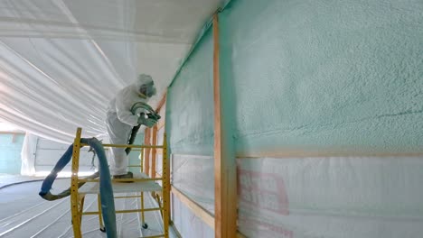 man in respirator and tyvek suit sprays closed cell foam insulation on exterior wall for new construction while standing on scaffolding