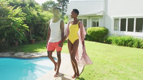 feliz pareja afroamericana caminando por la piscina tomados de la mano en un jardín soleado sonriendo