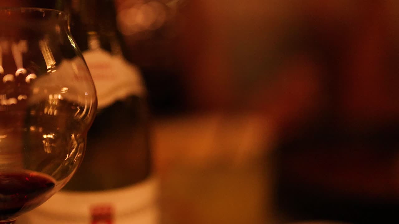A close-up of wine glasses and bottles in a dimly lit, warm-toned setting, capturing a relaxed, intimate atmosphere