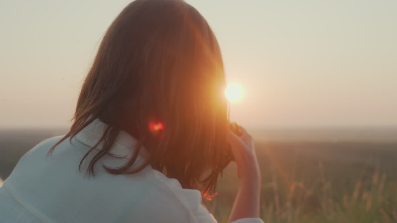 Lady enjoying peaceful outdoor picnic during sunset, picking up camera to take photo while sitting on grass beside glass of red wine, fresh fruit, flowers, and green landscape in warm golden light