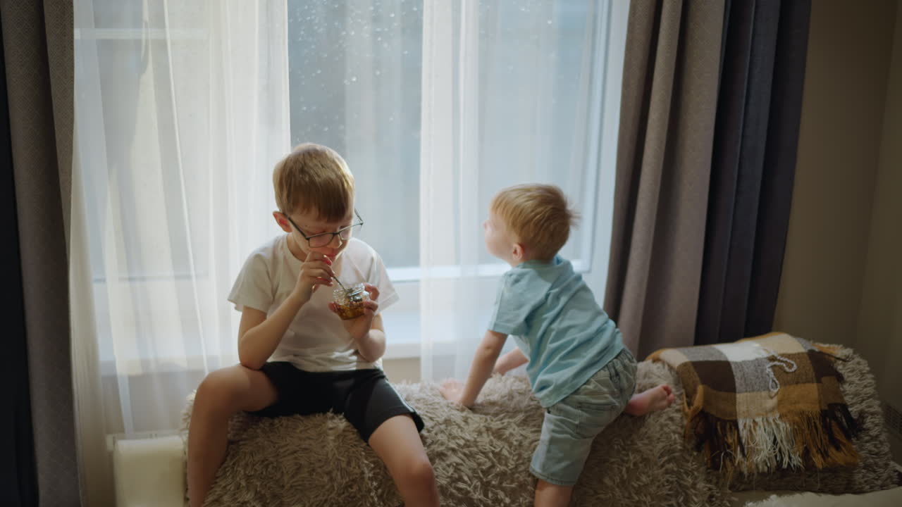Two young boys spending time indoors near window with sunlight, older boy wearing glasses eating from jar with spoon while younger boy in blue shirt climbs onto soft blanket surface, warm cozy atmosphere