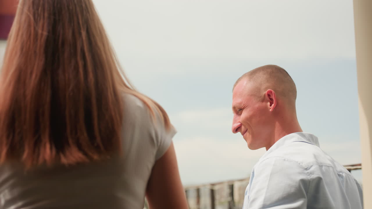 Rooftop date caucasian couple smiling on urban terrace, man looks out then turns to partner beside pink bubble seat. Warm sunlight bathes casual outfits, candid laughter and intimate gestures create