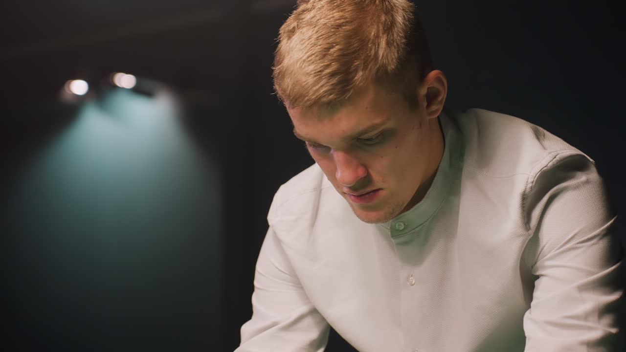 색 셔츠를 입은 젊은이의 클로즈업, 어두운 배경에 진정된 집중으로 깊이 초점을 맞추고, 오버헤드 빛은 그의 사려 깊은 표정을 강조하는 부드러운 빛을 만니다.