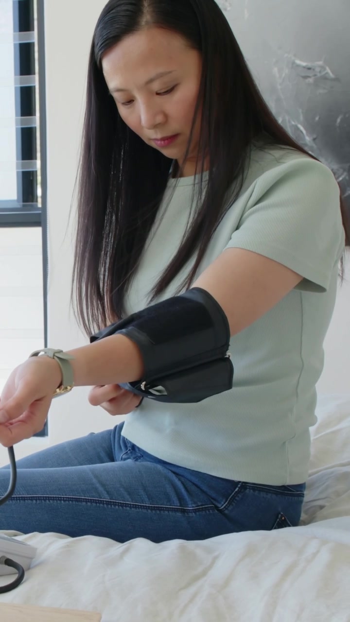 Video verticale: Donna con maglietta verde che prende il bracciale, lo fissa sul letto e misura la pressione sanguigna, con spazio per il testo.