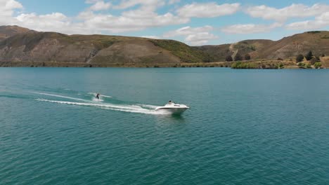 Cámara-Lenta:-Esquí-Acuático-Detrás-De-Un-Bote-En-El-Lago-Dunstan-Cerca-De-La-Represa-Clyde,-Otago-Central,-Nueva-Zelanda-Con-Montañas-Y-Nubes-En-El-Fondo