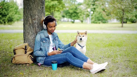 modern african american girl is listening to music with headphones and using smartphone relaxing in city park with pet dog, stroking and caressing the animal.