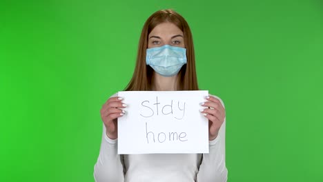 portrait of young girl in medical protective face mask looking at camera and holding a poster with the words stay home