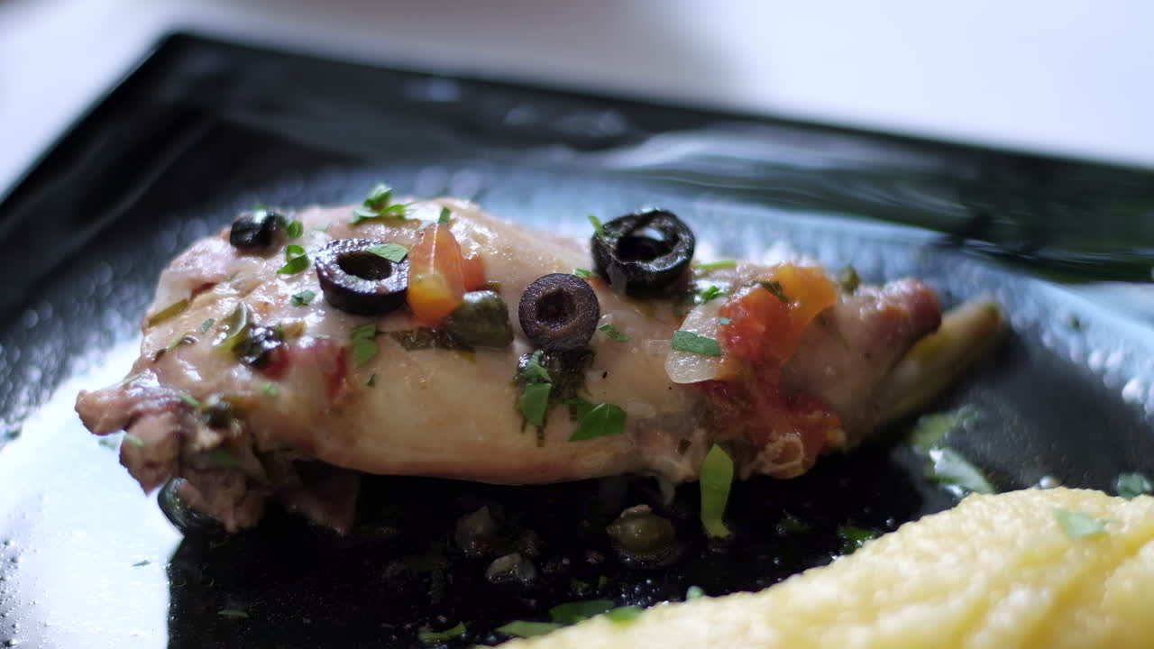 Close up of a stuffed chicken breast with black olives on top and mashed potatoes on the side in a black plate