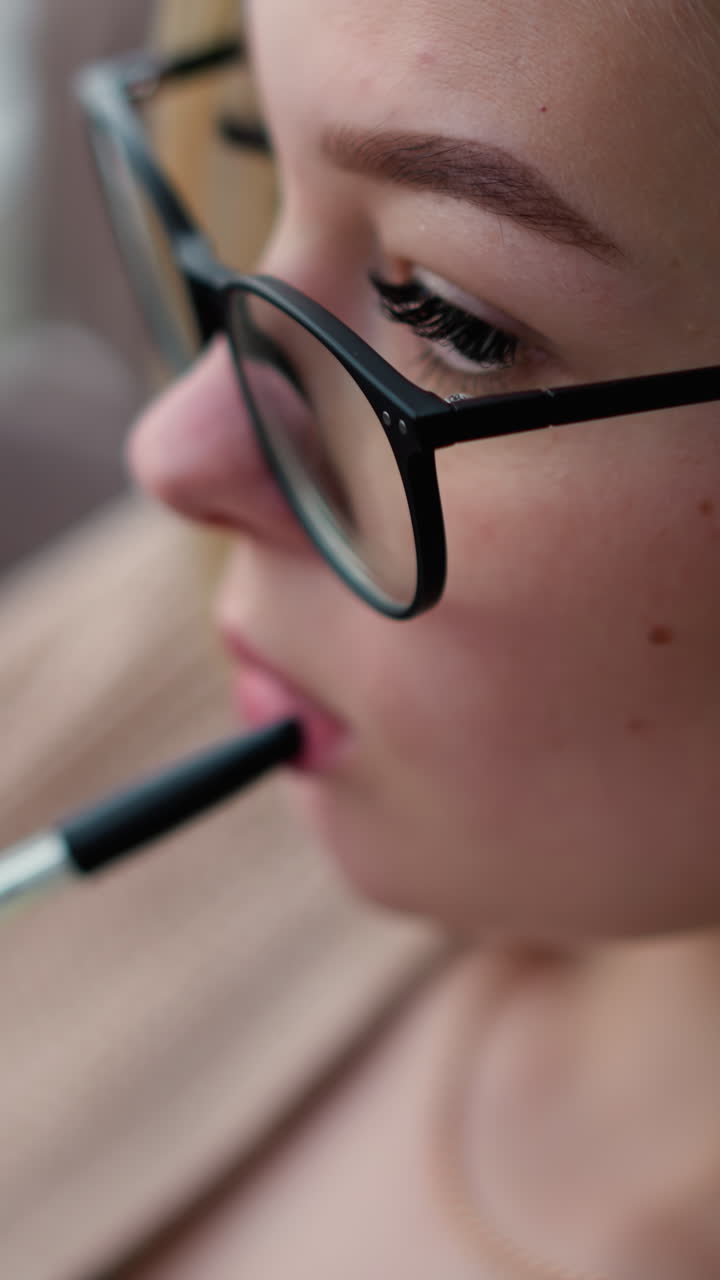 primer plano de una mujer blanca pensativa con la pluma suavemente presionada contra los labios, tocando pensativamente contra ellos mientras lleva gafas, fondo borroso que muestra contemplación o pensamiento profundo