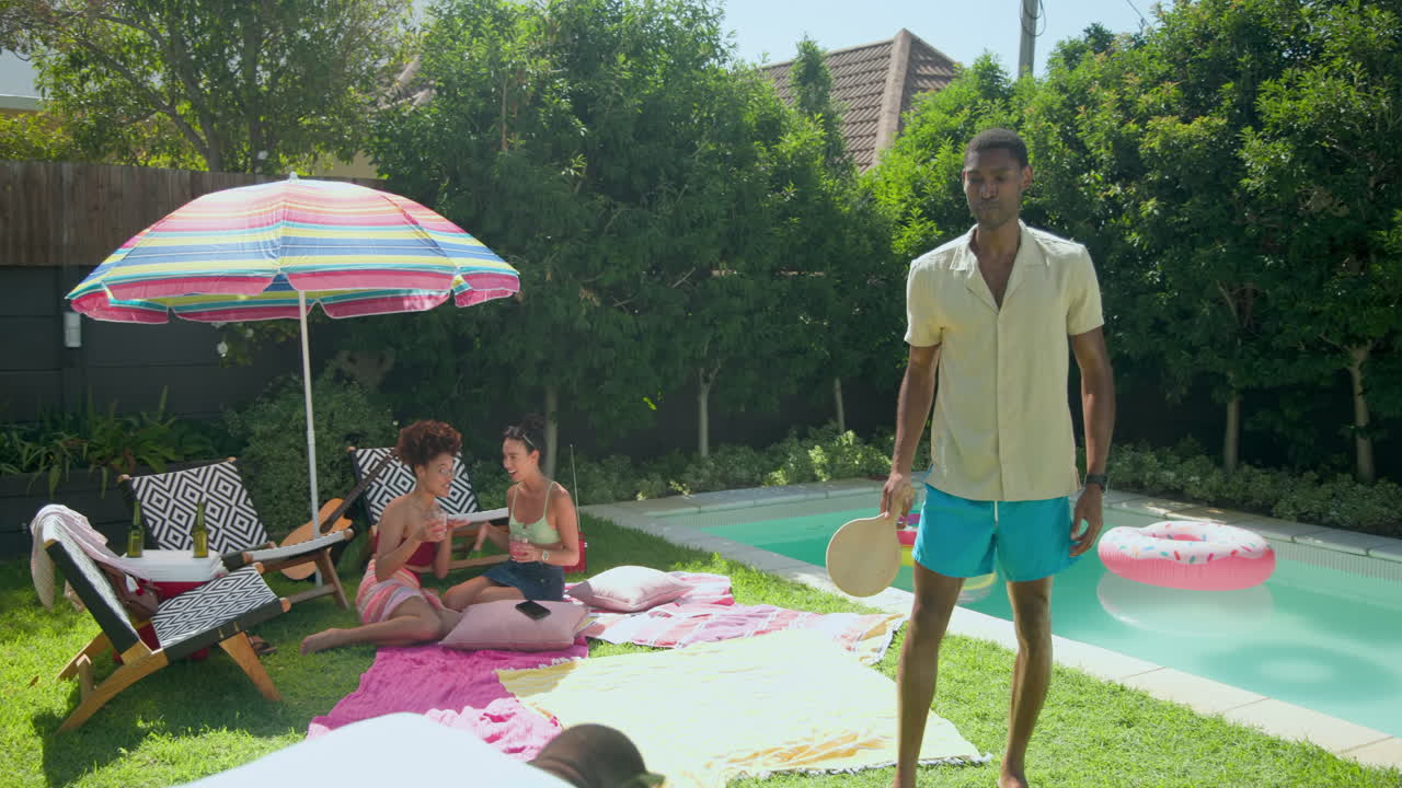 Diverse friends relaxing by pool under colorful umbrella, enjoying sunny day together