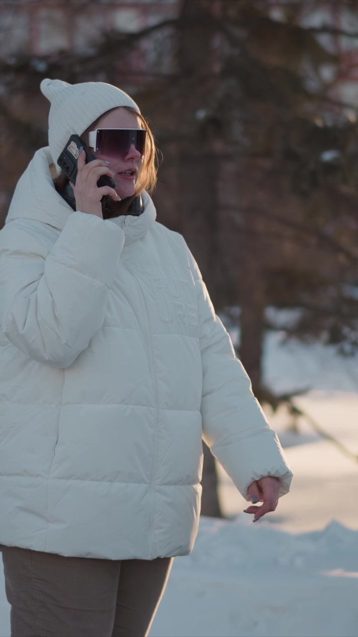 Woman wearing white puffer jacket and knit hat holds her smartphone engaged in conversation outdoors during winter. scene features soft golden sunlight filtering through trees with snow