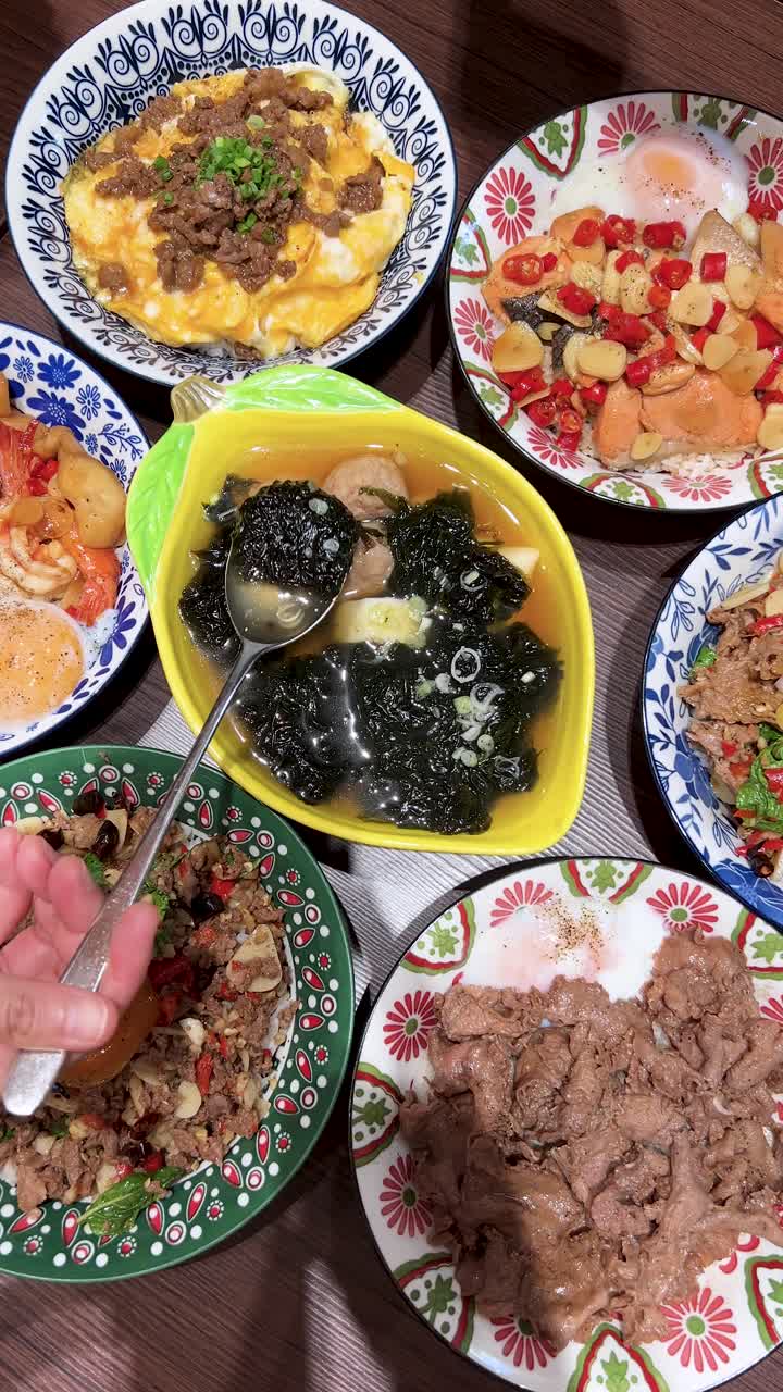 明るい室内照明の下、色鮮やかなタイ料理に囲まれた中央のボウルから、豚肉団子と海藻のスープを手でよそっている様子を真上から撮影。