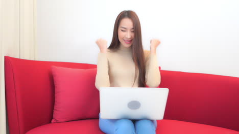 asian lady sitting with a wireless laptop on her knees on a red sofa in her living room using the device and tightening her fists with joy