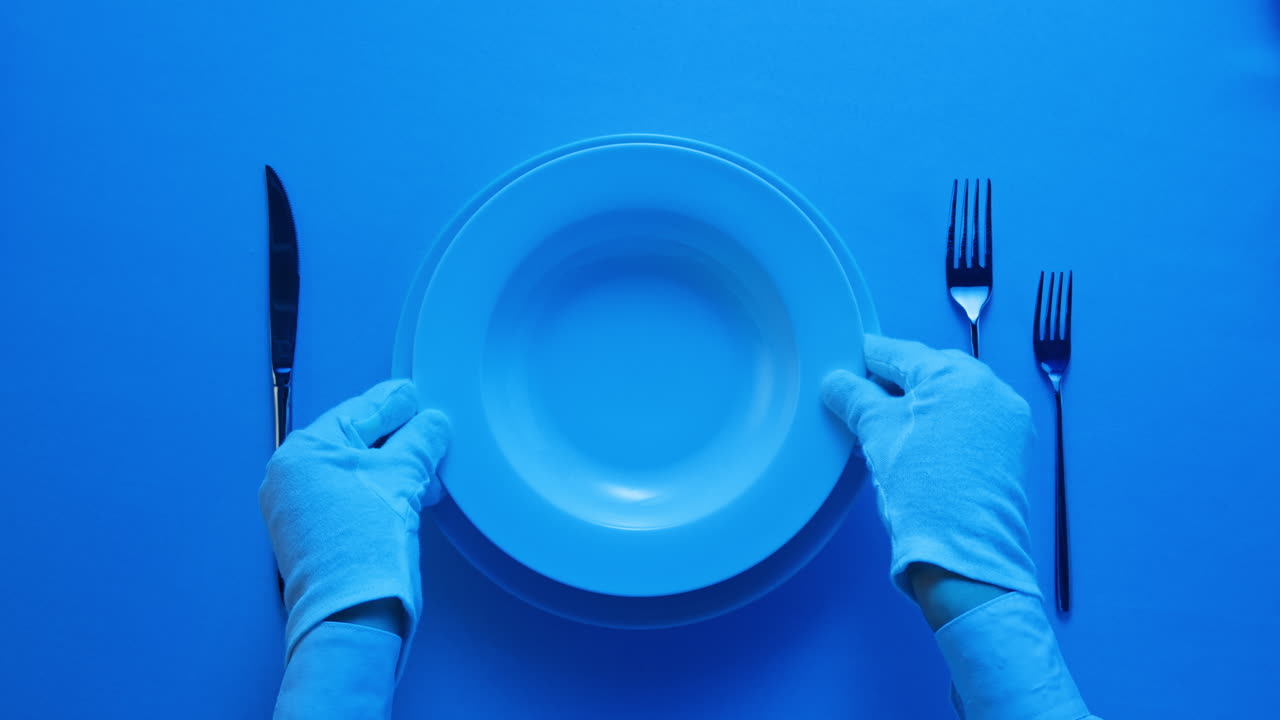 Minimalist table setting with white plate, fork, and knife arranged neatly, and a gloved hand adjusting cutlery.