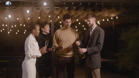 glücklicher mann, der eine flasche champagner öffnet und mit seinen fröhlichen freunden das neue jahr feiert