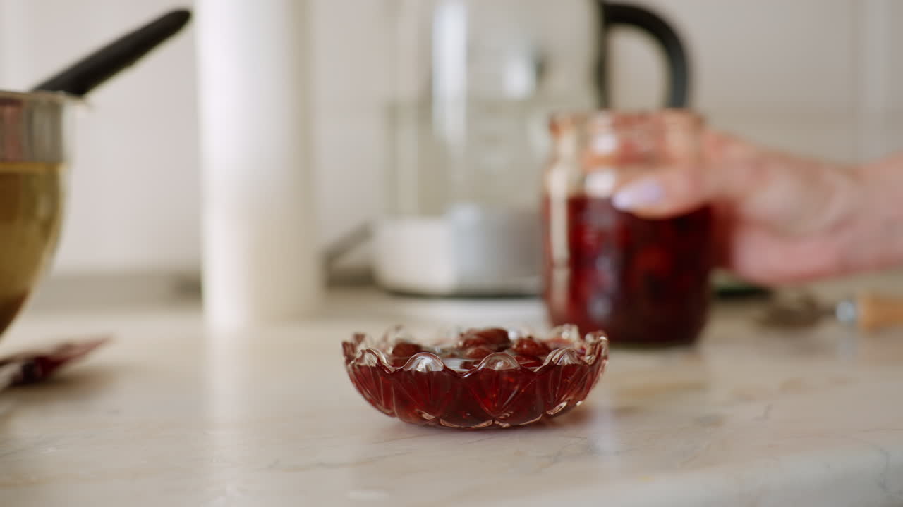 Red fruit jam dropped from hand-held jar into decorative glass dish on clean kitchen counter, with blurred kettle and bowl in background, showing detail of preserved fruit being served