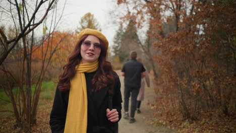 lady with red curly hair walks through forest path with autumn foliage, smiling, carrying black bag, as two persons pass by in opposite direction