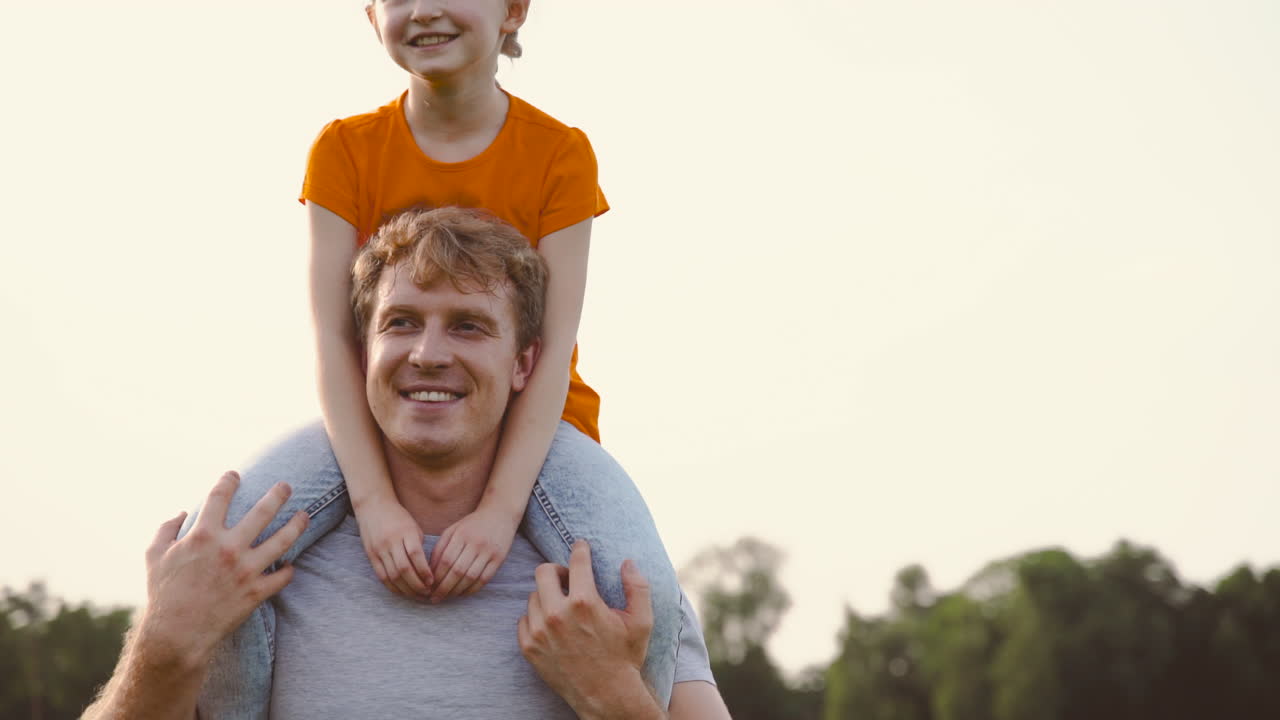걷고 그녀와 이야기하는 동안 공원에서 어깨에 그의 웃는 딸을 들고 행복한 아버지