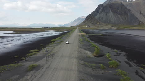 Aerial-of-4x4-car-approaching-on-gravel-black-road-between-water-from-both-sides-and-with-black-mountains-and-cloudy-sky-in-the-background