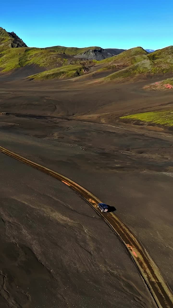 Vista aerea verticale con drone di un fuoristrada 4x4 solitario che segue una strada sterrata curva attraverso gli altipiani vulcanici islandesi, con pianure di cenere nera e colline di riolite verde sotto un cielo azzurro limpido