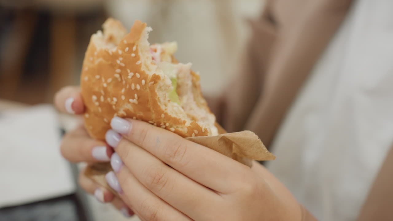 Primer plano de una mujer mordiendo una hamburguesa en la cafetería de un centro comercial; mujer caucásica con manicura disfrutando de una hamburguesa en interiores; primer plano de una mujer elegante saboreando una hamburguesa con manicura en la cafetería de un centro comercial.