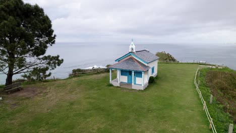 Small-blue-and-white-church-next-to-the-sea,-Ermita-de-la-Regalina,-above-the-steep-cliffs-on-the-coast-of-Asturias,-Spain